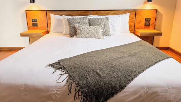 Una cama bien hecha con edredón blanco, una manta gris y varios cojines estampados. Hay dos mesitas de noche con lámparas y un cabecero de madera.