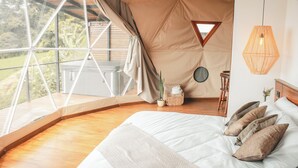 Un dormitorio con un ventanal grande, una cama con almohadas, una lámpara colgante y una pequeña mesa redonda con una planta.