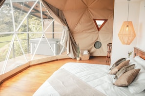 A bedroom with a large window, a bed with pillows, a hanging light fixture, and a small round table with a plant. - Ananta Forest (Monteverde)