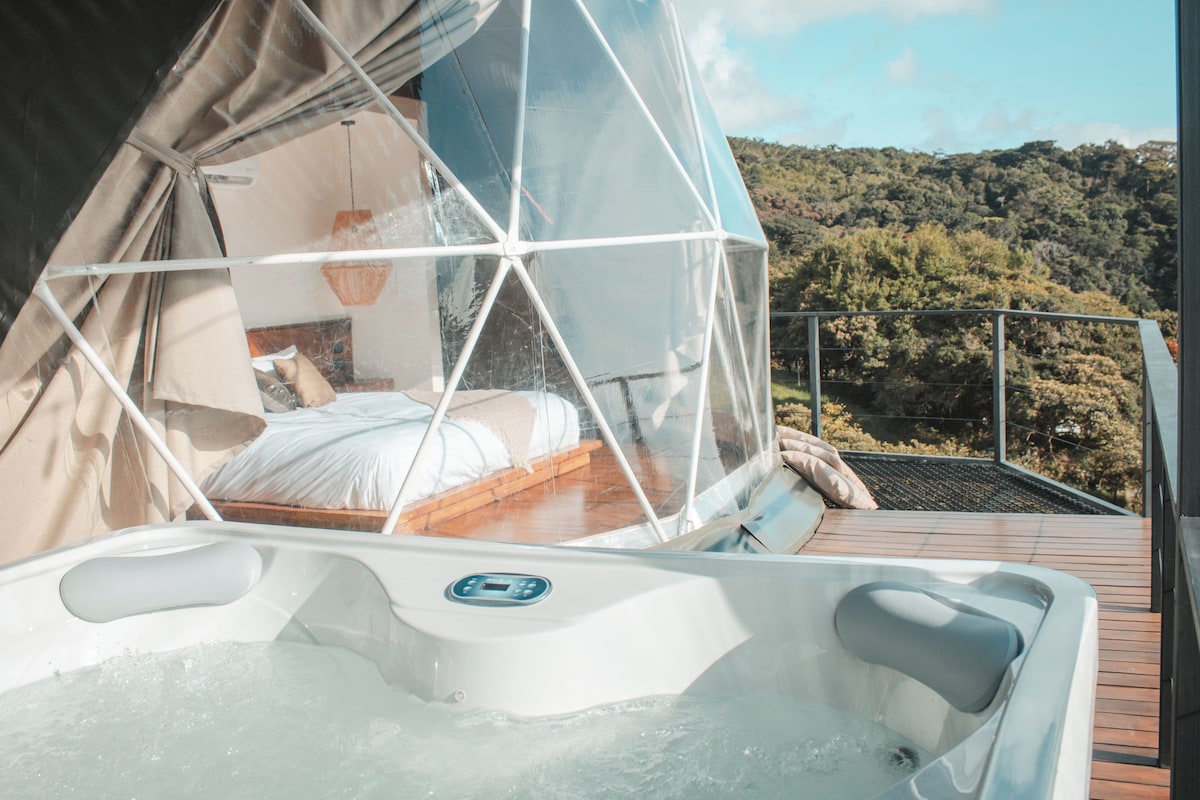 A hot tub with a wooden deck and a bed under a transparent canopy.