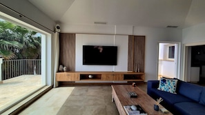 Una sala moderna con una TV de pantalla plana montada en una pared de madera, un sofá azul, una mesa de centro y un puerta corrediza de vidrio que da a un balcón.