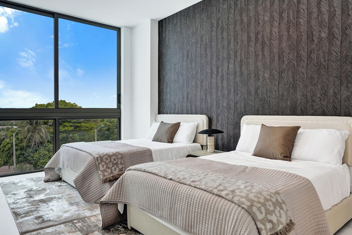A modern hotel room with two beds, large windows, a view of greenery, and a textured wall.