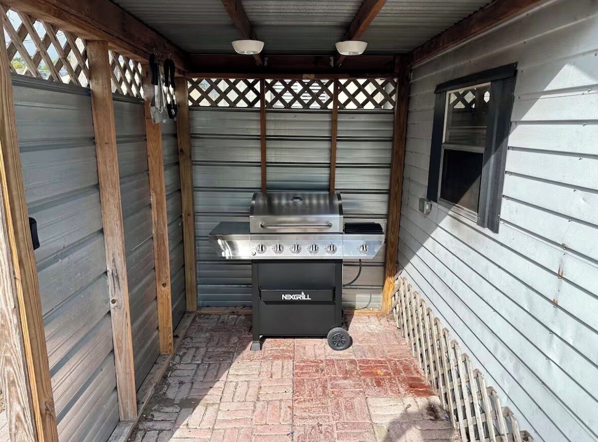 A barbecue grill with four burners and a side burner, located in a covered outdoor area with a wooden lattice ceiling and a window on the right.