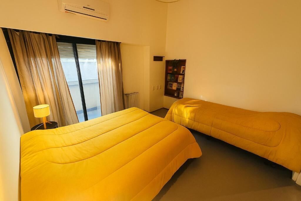 A hotel room with two beds, a window with curtains, a radiator, and a bookshelf.
