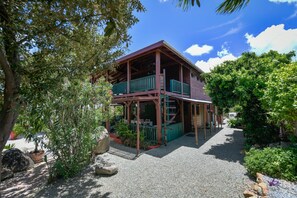 A two-story wooden house with a balcony, surrounded by greenery and a gravel driveway. - Villa  Luna LLena located in the Jamanota hills of Aruba's Arikok National park (Santa Cruz)