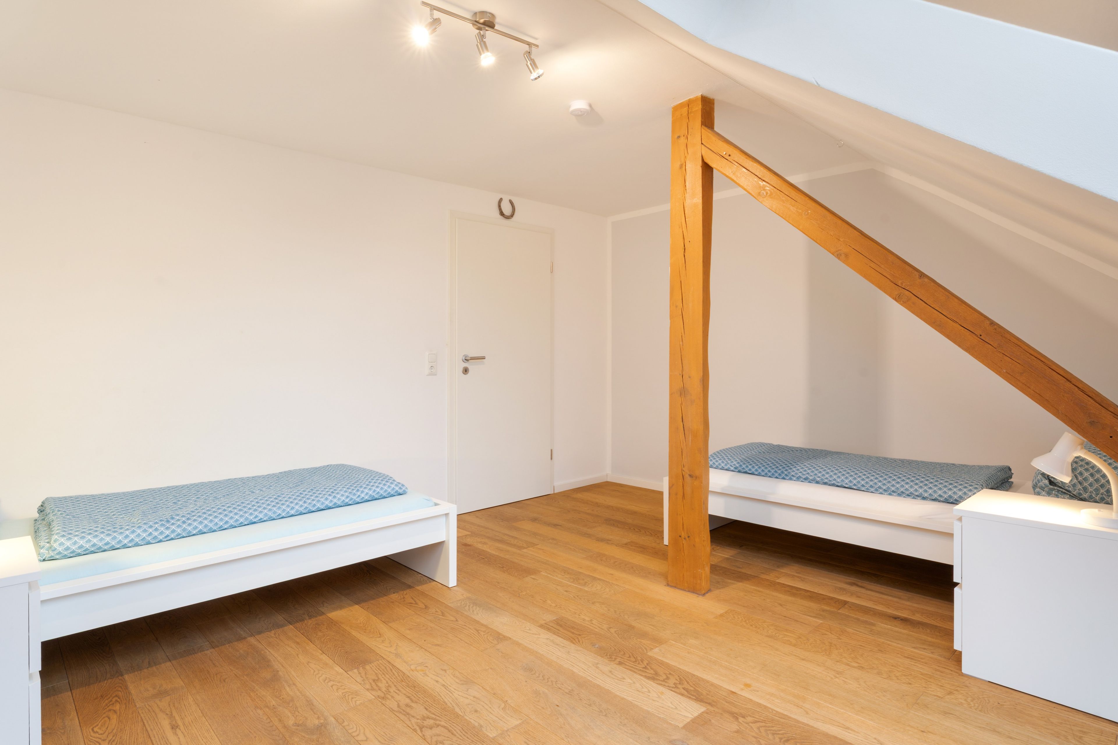 Een kleine, goed verlichte kamer met een schuine kap, twee bedden met blauw beddengoed, een witte deur en een houten balkenstructuur.