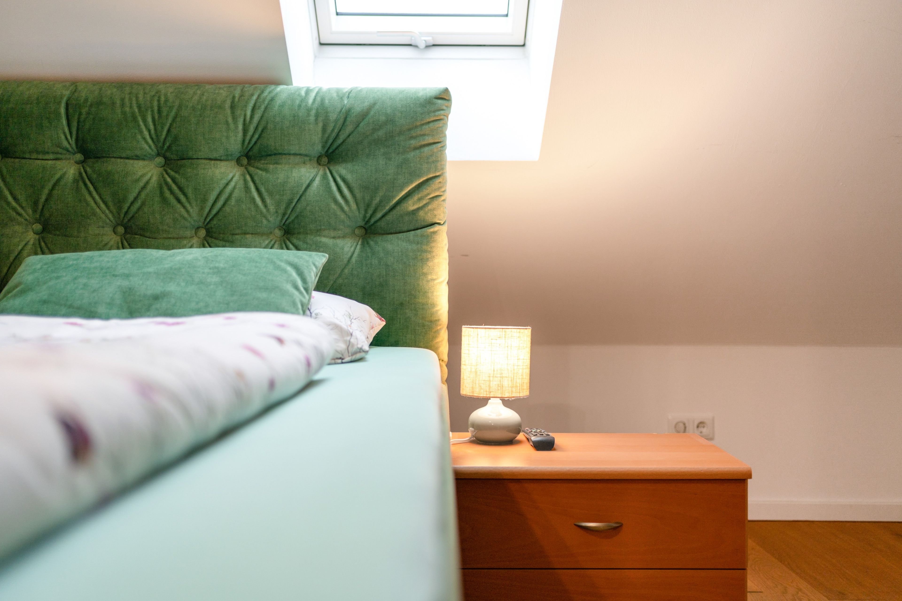 Un dormitorio con una cama, una mesita de noche con lámpara y una ventana sobre la cama.