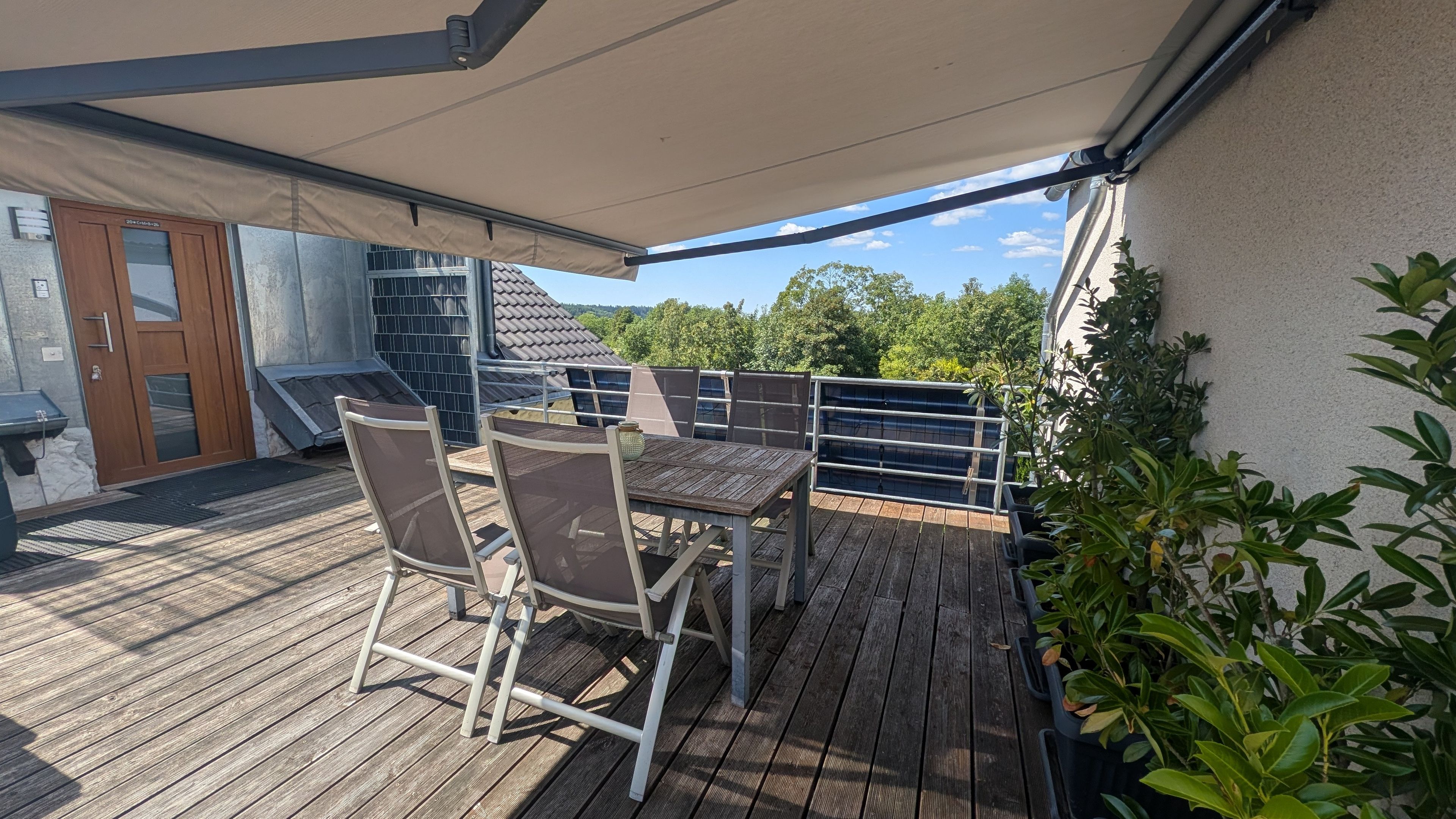 Une terrasse en bois avec une table et des chaises, un espace abrité et une vue sur des arbres et un bâtiment.