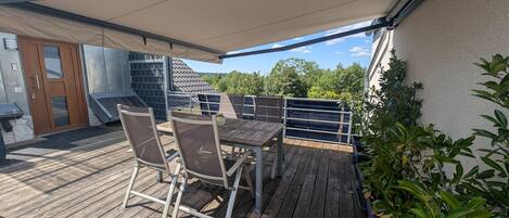 Une terrasse en bois avec une table et des chaises, un espace abrité et une vue sur des arbres et un bâtiment.