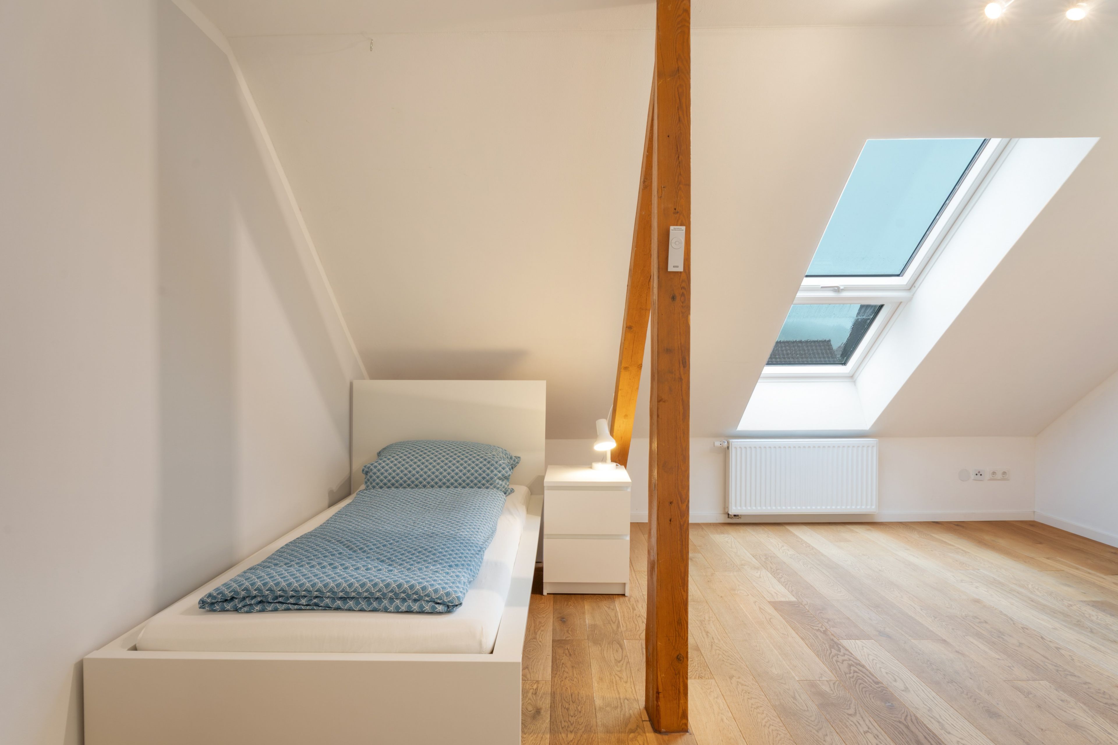 Una habitación pequeña y luminosa con una cama individual, una mesita de noche y un tragaluz.
