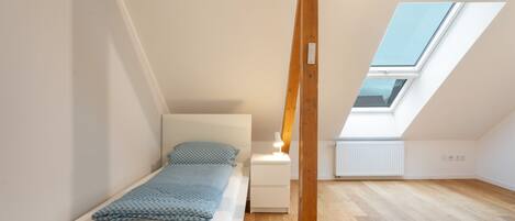 A small, well-lit room with a single bed, a nightstand, and a skylight.