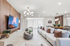 Uma sala de estar moderna com televisão de tela plana, uma mesa de centro redonda, um conjunto de sofás e uma área de jantar com mesa e cadeiras de madeira.