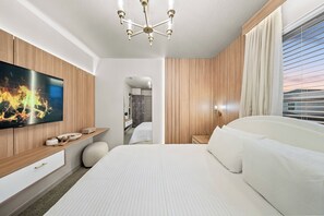 Ein modernes Hotelzimmer mit einem großen Bett, einem flachbildfernseher an einer Holzvertäfelung, einem Nachttisch mit Lampe und Blick ins Freie durch das Fenster.