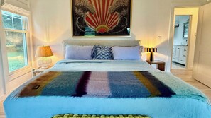 A bedroom with a bed, a painting above the bed, a lamp on the nightstand, and a view of a hallway.