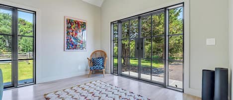 A spacious room with large windows, a wooden floor, a colorful rug, a chair, and a framed painting.