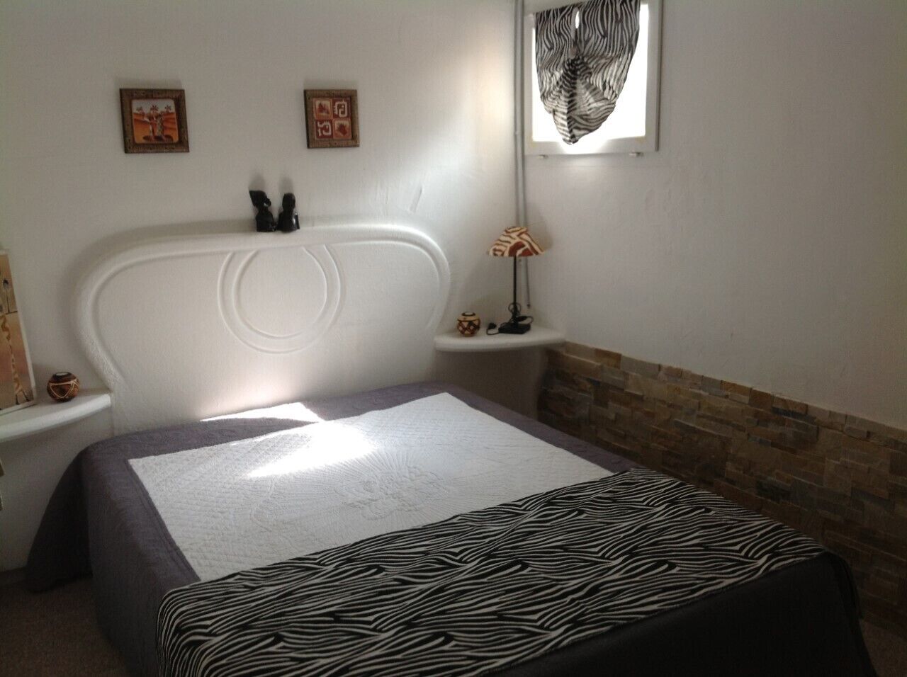 A bedroom with a large bed, a nightstand, a wall-mounted lamp, and a window with a zebra-patterned curtain.