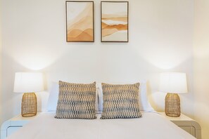 Un lit bien fait, agrémenté de deux coussins à motifs, de deux lampes fixées au mur et de deux œuvres d’art encadrées au-dessus.