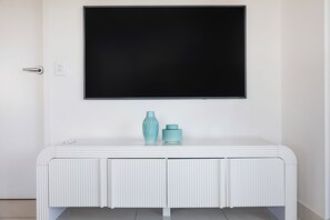 Une console blanche ornée de deux vases décoratifs, un téléviseur noir fixé au-dessus, et une porte dotée d’une poignée argentée.