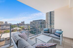 A balcony with a sofa and a table, overlooking a cityscape with modern buildings. - Capital Panorama Central City Escape w Pool and Gym (Darwin)