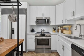 A modern kitchen with white cabinetry, stainless steel appliances, a wooden table, and a herringbone-patterned backsplash. - The Banana Cabana - Cozy Cottage (West Palm Beach)