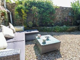 Un espacio al aire libre con sillones de mimbre, una mesita baja con dos tazas y una pared de piedra cubierta de plantas trepadoras.