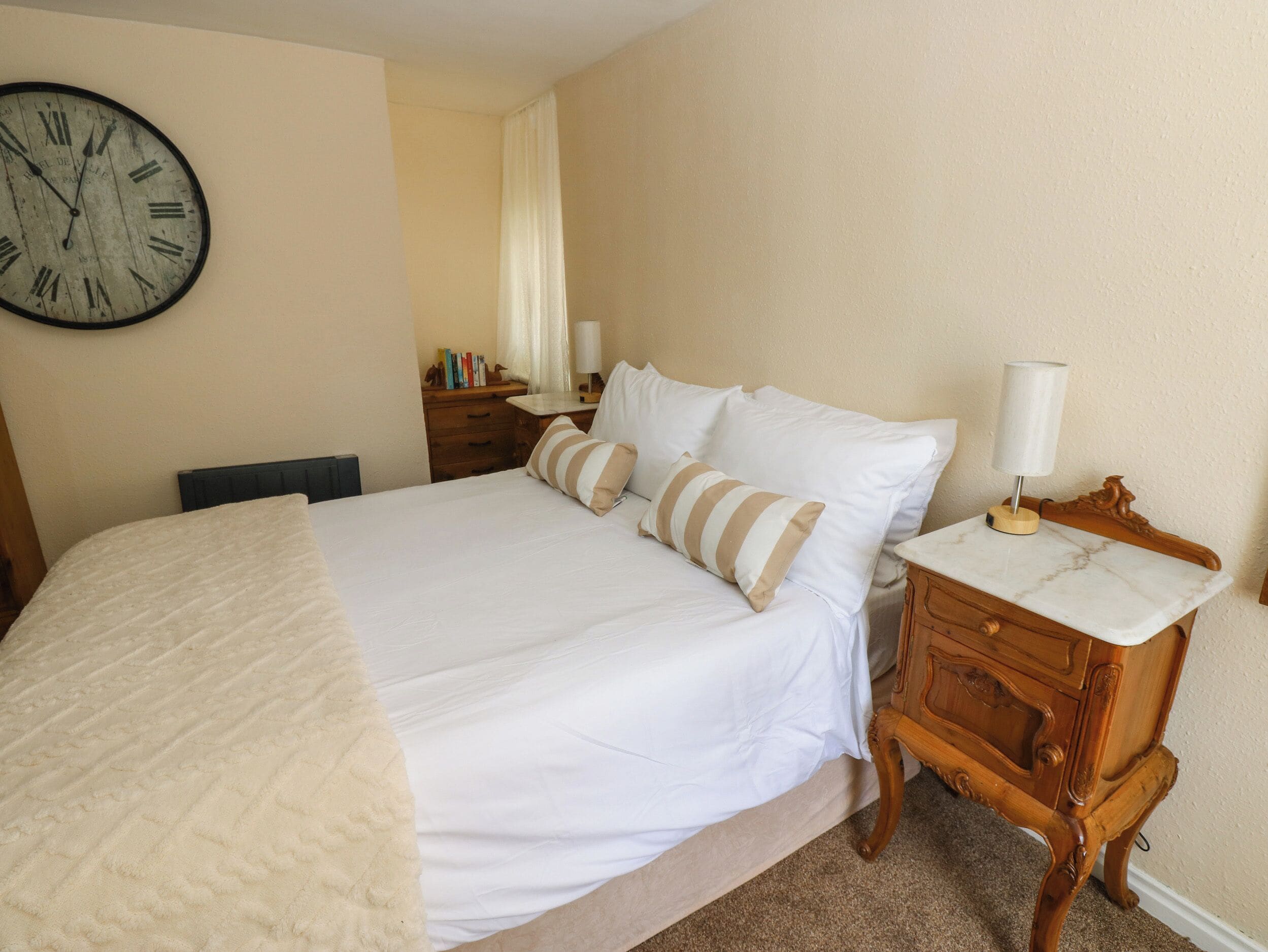 A neatly made bed with a white comforter and pillows, a wooden nightstand with a lamp, and a wall clock.