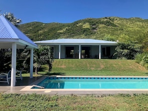 A pool with a clear blue water, a covered area with chairs, and a house with a white exterior and blue roof. - Villa Sunrise Indigo – Spacious Retreat with Gorgeous Sea Views (Cul-de-Sac)