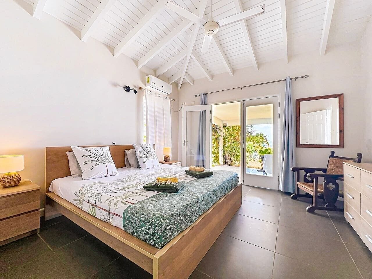 A spacious bedroom with a large bed, a wooden headboard, a nightstand with a lamp, a dresser, and a sliding glass door leading to a balcony.