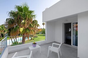 Un balcon avec des chaises blanches, une petite table sur laquelle est posé un pot de fleurs, et une vue sur une piscine et des palmiers.