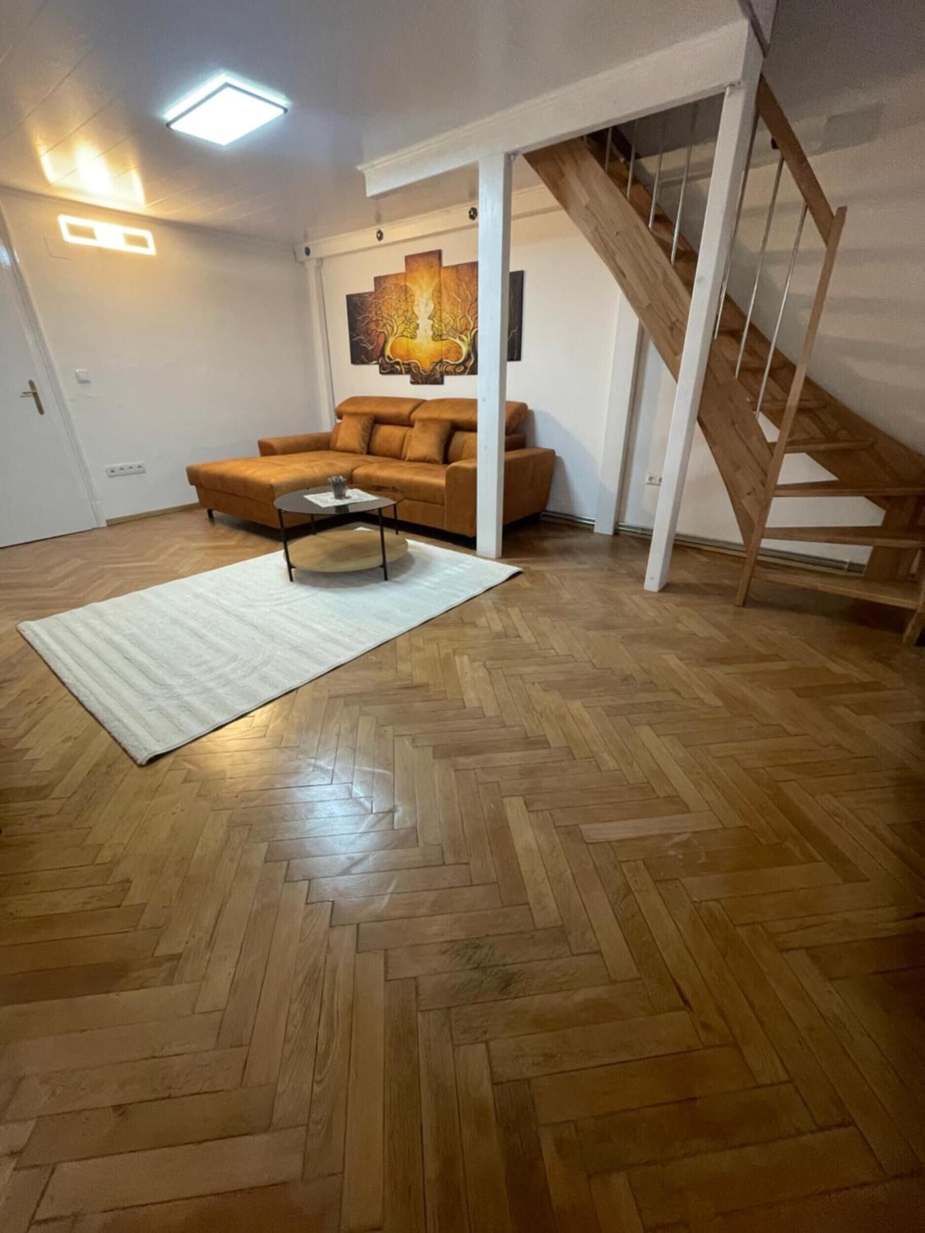 A living room with a herringbone wooden floor, a brown sofa, a coffee table, and a painting on the wall.