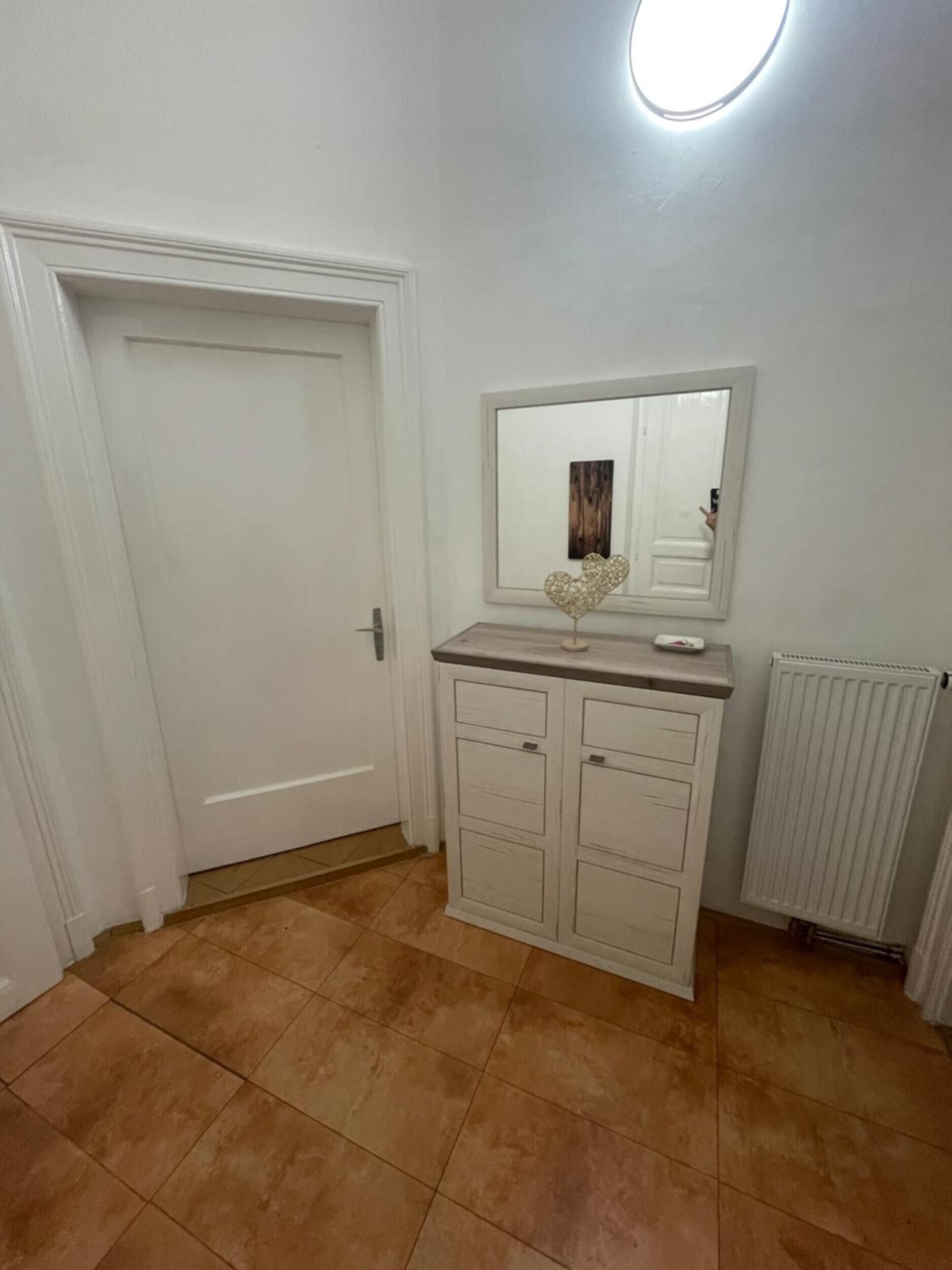 Una habitación con una puerta blanca, un armario blanco con cajones, un espejo sobre el armario y un radiador en la pared.