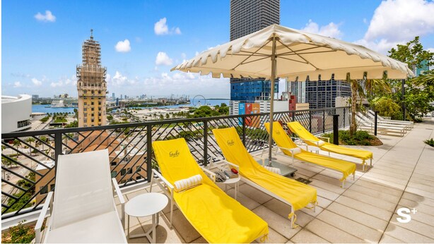 A rooftop lounge with yellow sunbeds and a large umbrella, overlooking a cityscape with a tall building and a tower. - Cozy Miami Studio - Pool & Gym - Views (Miami)
