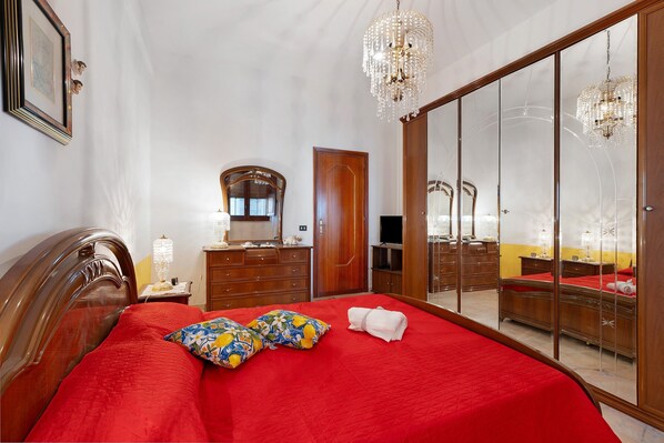 A hotel room with a large bed, a wardrobe with a mirror, a chandelier, and a television.
