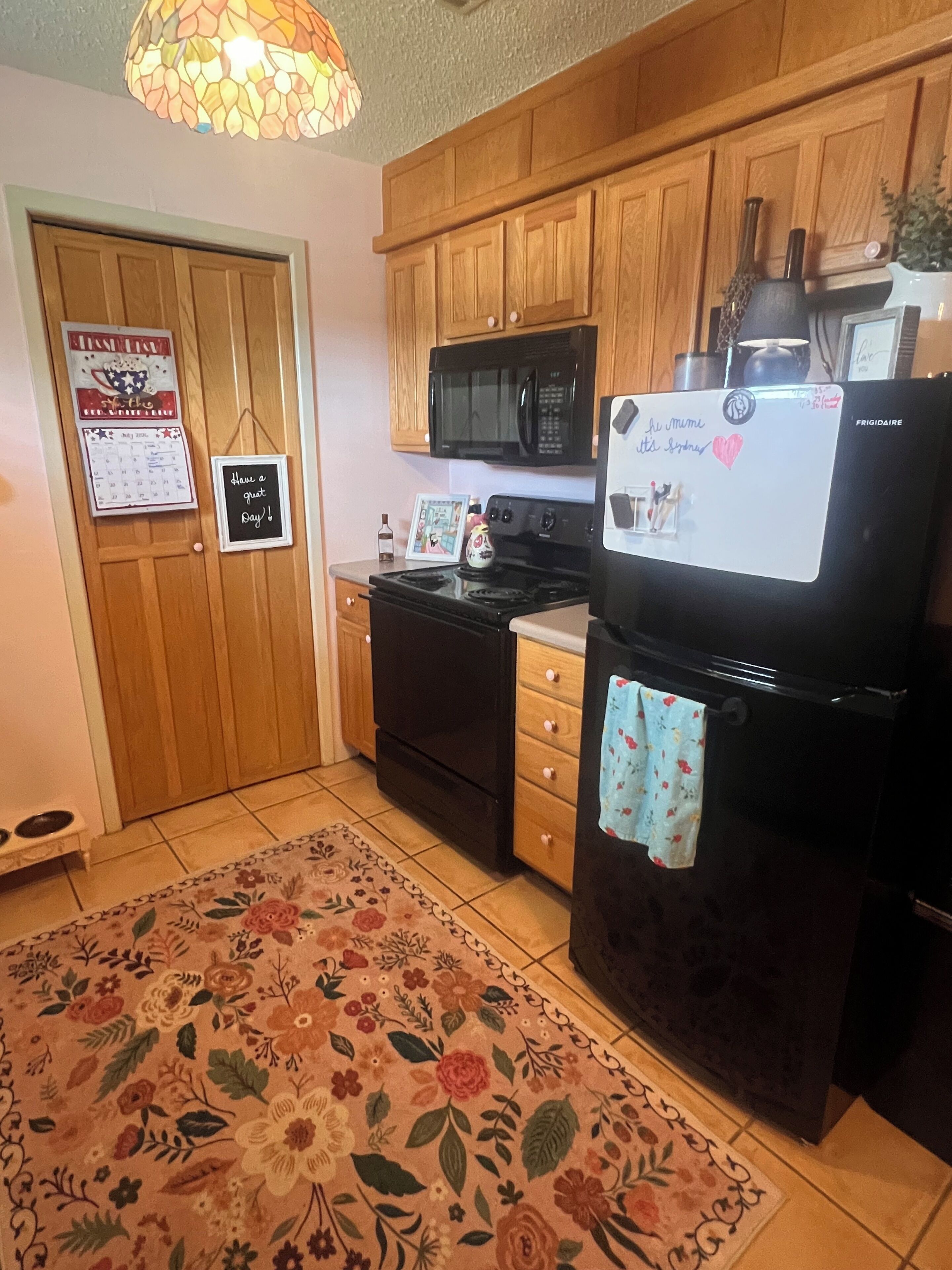 Una cocina con armarios de madera, un refrigerador negro, un microondas y una estufa. Hay una alfombra estampada en el suelo y una lámpara colgante.