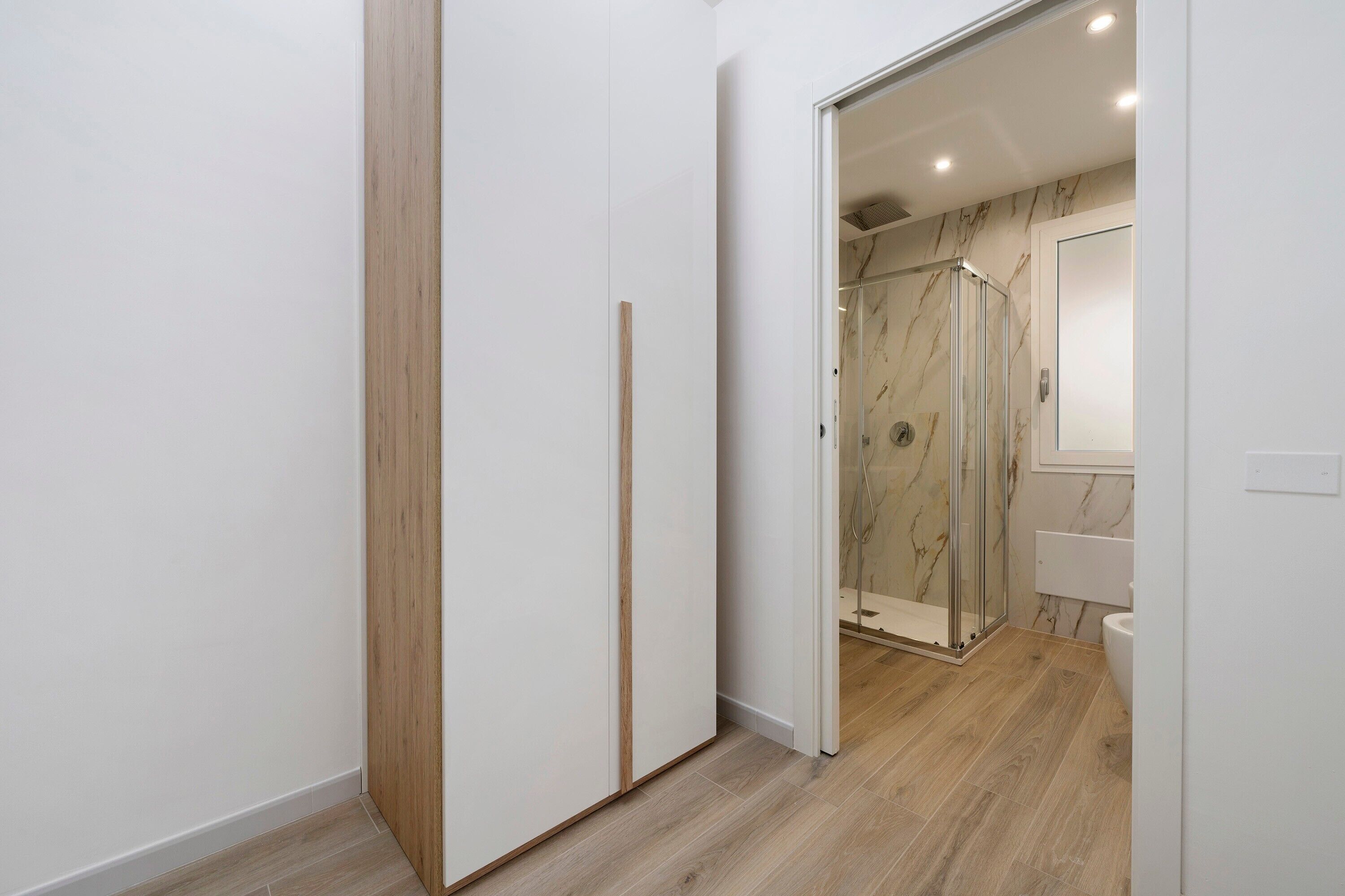 Een hal met een houten vloer, een glazen douchecabine en een witte deur die leidt naar een badkamer met een marmeren wand en een witte wastafel.