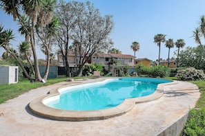 A swimming pool with a curved edge, surrounded by a concrete deck. There are palm trees and a building with a red roof in the background. - Villa Matilde Domo Boutique – Suite Pietra Daniele (Torre degli Ulivi)