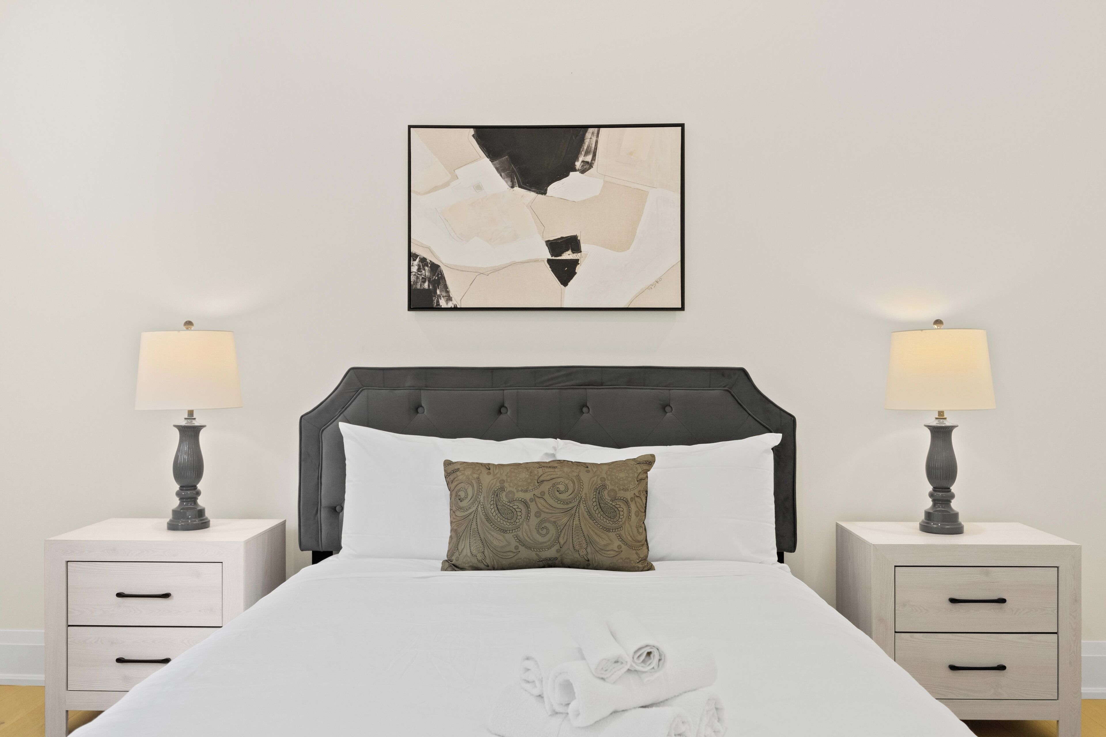 A neatly made bed with a headboard, two bedside tables with lamps, a pillow, and a wall-mounted abstract painting.