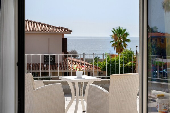 A balcony with wicker furniture and a potted plant, overlooking a coastal area with buildings and a palm tree. - Villa Matilde Domo Boutique - Master Suite with Sea View (Torre degli Ulivi)