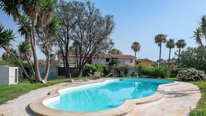 A swimming pool with a curved edge, surrounded by a concrete deck. There are palm trees and a building with a red roof in the background. - Villa Matilde Domo Boutique - Master Suite with Sea View (Torre degli Ulivi)