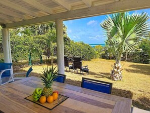 A covered outdoor dining area with a table set for two, surrounded by chairs, and a view of the ocean and tropical vegetation. - Villa Sunrise Blue – Peaceful Retreat with Bay Views in Cul-de-Sac (Cul-de-Sac)
