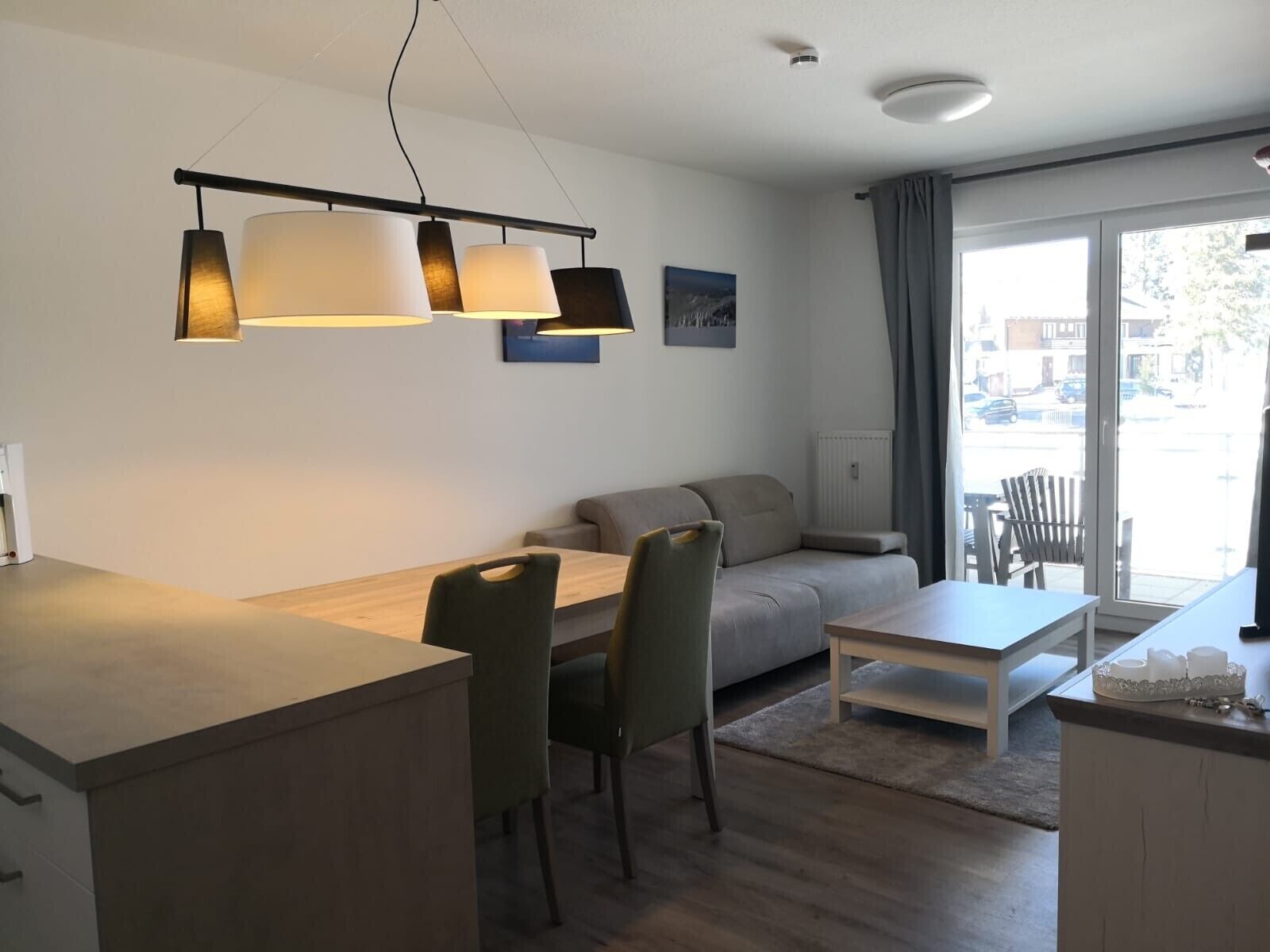 Een moderne woonkamer met een bank, eettafel, stoelen en een salontafel. Er is een groot raam met gordijnen, een radiator en een plafondlamp.