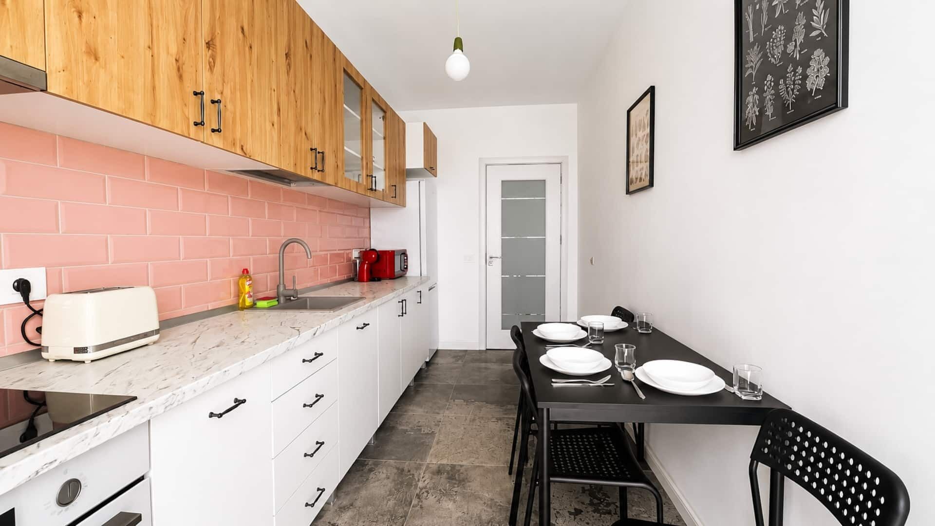Een kleine keuken met houten kastjes, een wit aanrecht, een spoelbak en een eethoek met een tafel gedekt voor twee personen.