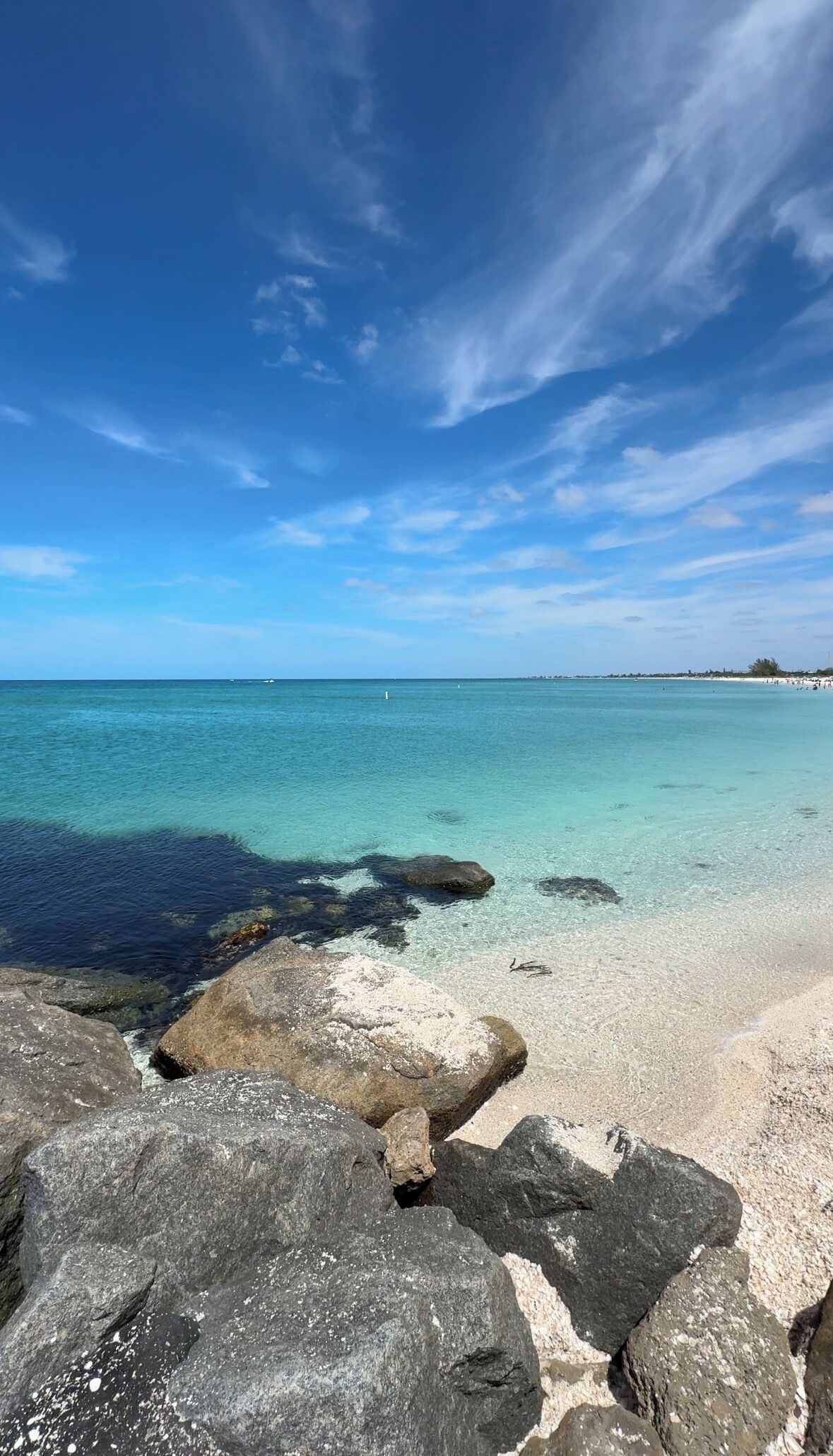 Une plage bordée d’une eau turquoise et limpide, avec un premier plan rocheux et une côte lointaine.