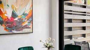Ett modernt matområde med ett glasbord, grönklädda stolar, en vas med vita blommor och en färgstark, abstrakt målning på väggen.