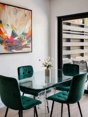 Ein moderner Essbereich mit einem Glastisch, grünen Polsterstühlen, einer Vase mit weißen Blumen und einem farbenfrohen abstrakten Gemälde an der Wand.