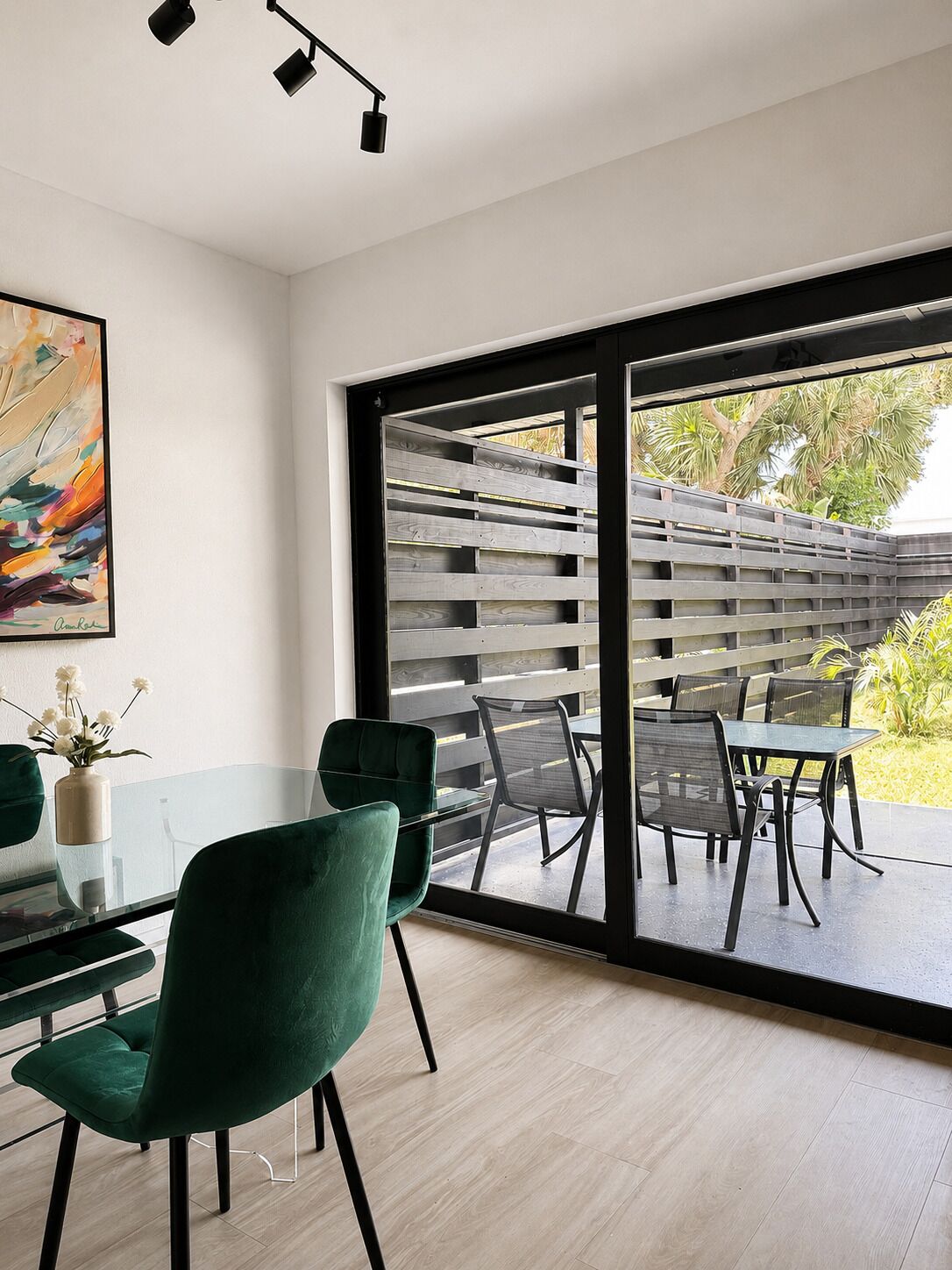 Un comedor moderno con una mesa de vidrio, sillas tapizadas en verde y vistas a un patio exterior a través de puertas correderas grandes.