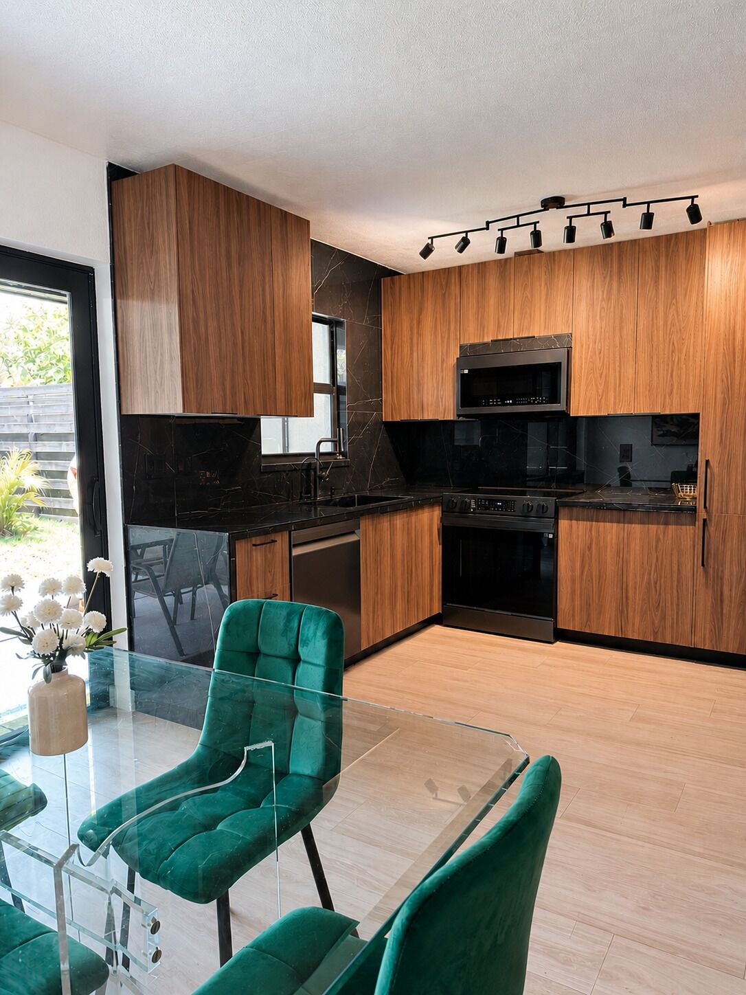 Cozinha moderna com armários de madeira, bancada preta e eletrodomésticos de aço inoxidável. Há uma mesa de jantar de vidro com cadeiras estofadas em verde.