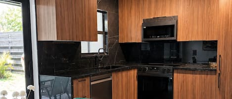 Una cocina moderna con armarios de madera, encimera negra y electrodomésticos de acero inoxidable. Hay una mesa de comedor de vidrio con sillas tapizadas de color verde.