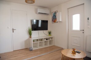 A room with a flat-screen TV mounted on the wall, a white console table with potted plants, a round wooden table with a vase and remote, and a door with a window. - The Little Farmhouse in Amiens - 1-Bedroom Apartment - Air-Conditioned! (Amiens)