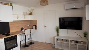 Una cocina moderna con horno empotrado, comedor con dos taburetes tipo bar, televisor de pantalla plana y un estante blanco con plantas.